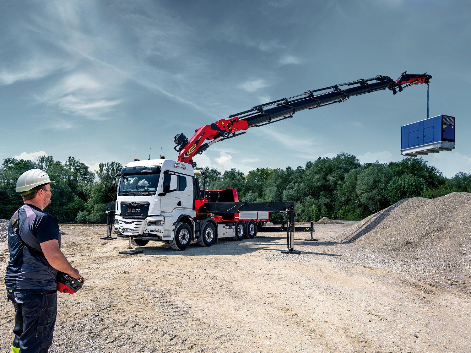 Truck-mounted knuckle boom crane lifting a blue industrial unit over a gravel site while an operator controls the crane remotely.